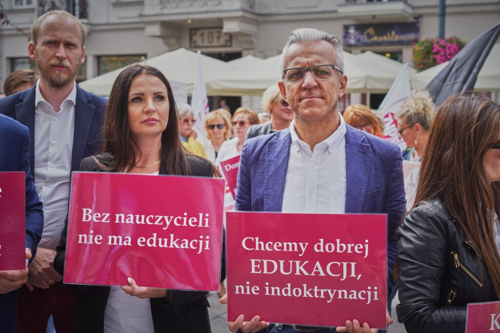 Two people at a demonstration holding signs reading “No education without teachers” and “We want good education, not indoctrination”
